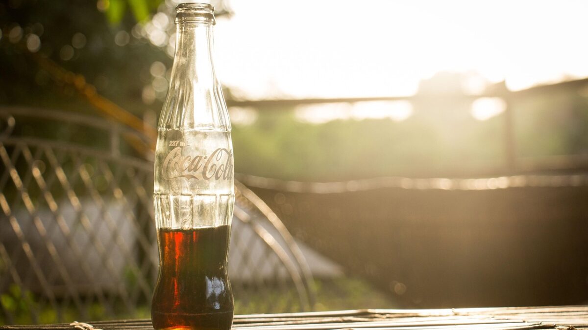 Une bouteille en verre Coca-Cola de 237 mL, à moitié remplie d'un liquide brun, posée sur une table en bois, à contre-jour d'un coucher de soleil, avec une clôture et de la verdure floues en arrière-plan.