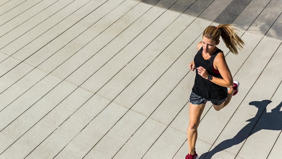 Une femme en tenue de running noire, baskets rouges et queue de cheval court sur une terrasse en bois clair ensoleillée, une montre connectée au poignet, vue en plongée.