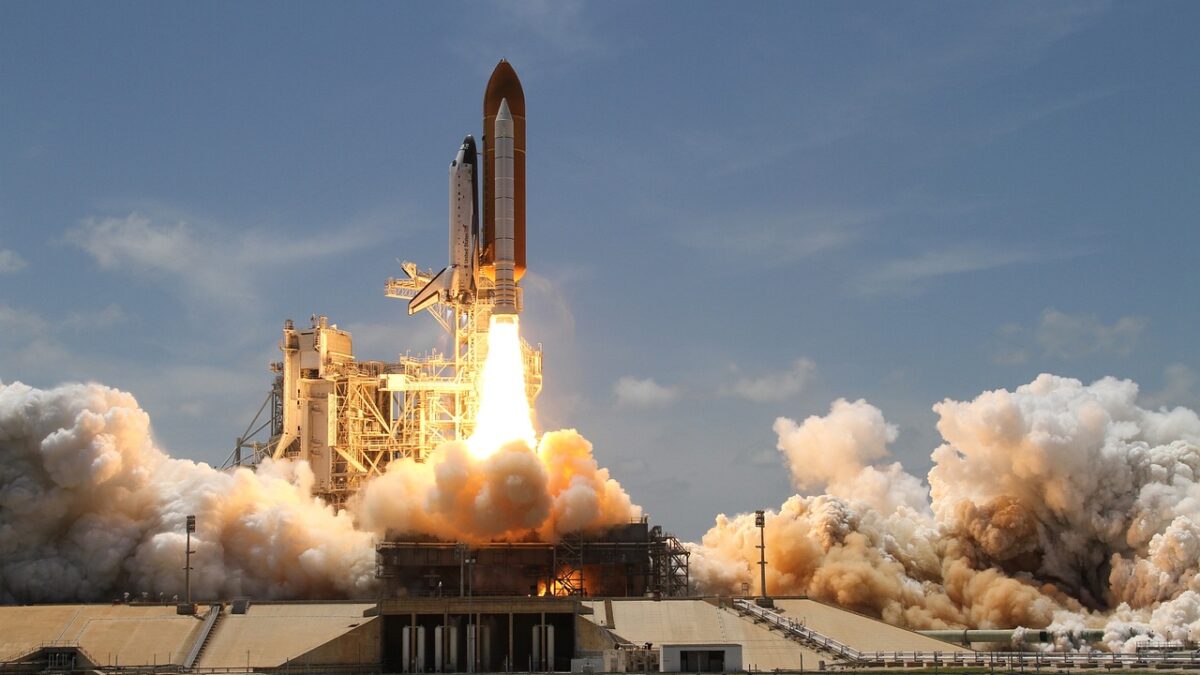 Lancement d'une navette spatiale depuis un pas de tir, avec son réservoir externe orange et ses propulseurs d'appoint, entouré d'un nuage de fumée et de flammes sous un ciel bleu.
