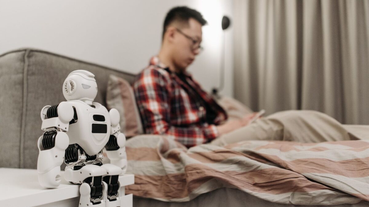 Petit robot blanc sur table de chevet, homme en chemise à carreaux allongé sur le lit regardant son téléphone.