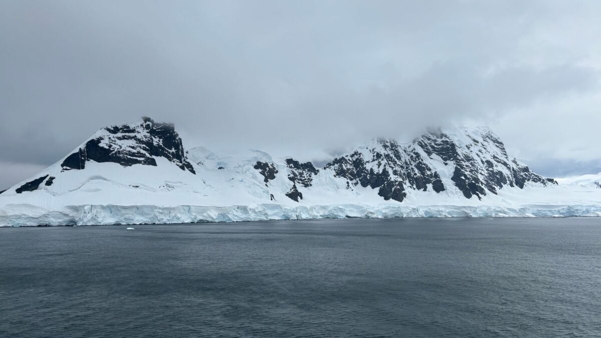 Montagnes enneigées et glace antarctique au bord d'une mer sombre sous un ciel nuageux.
