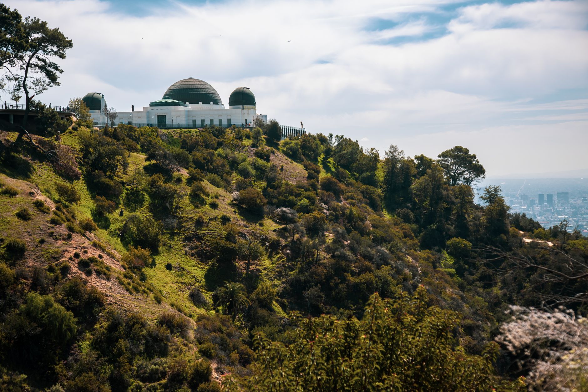Griffith Observatory sits on a green, brush-covered hillside overlooking the city in the distance.