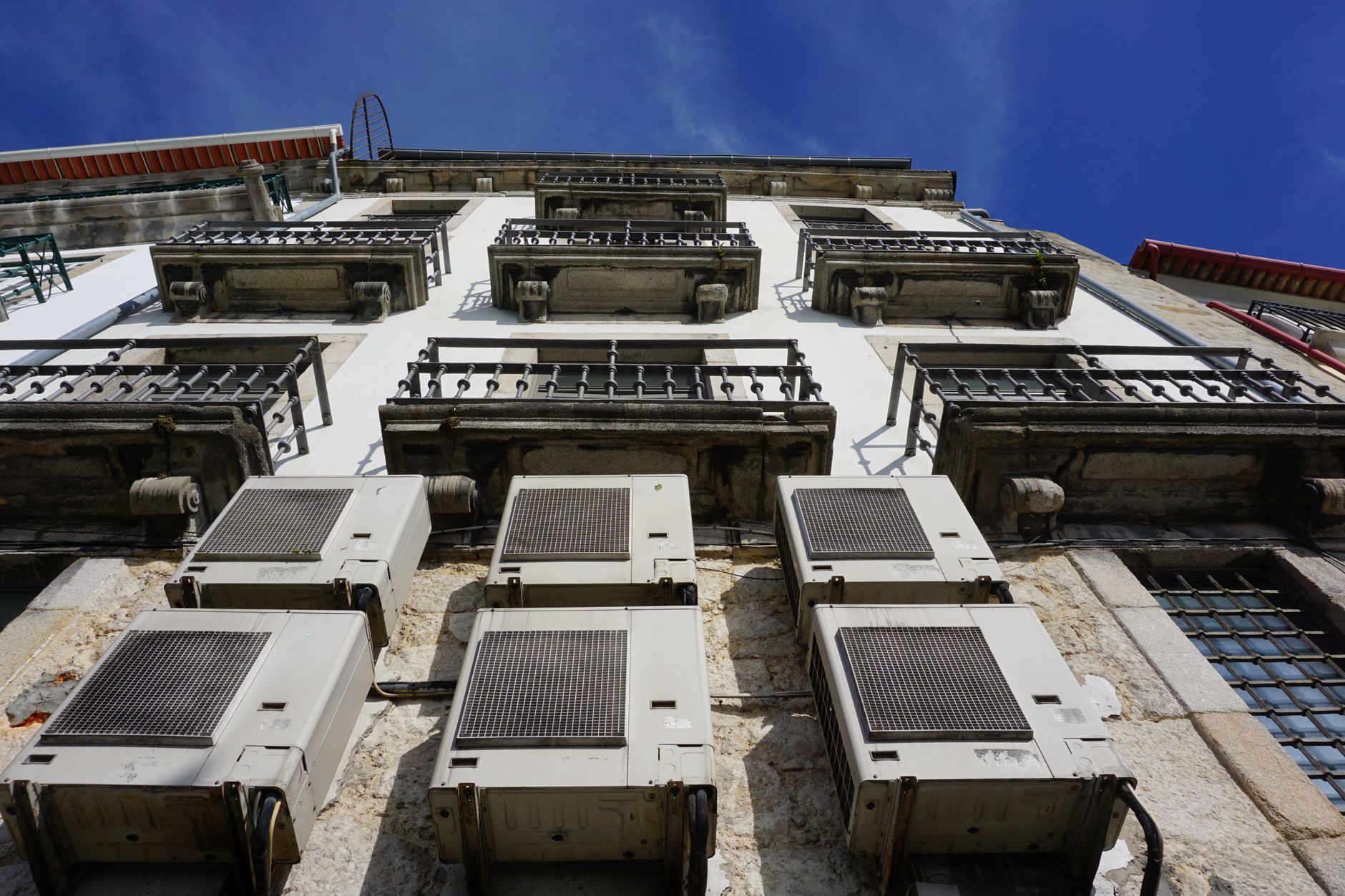 Six outdoor air conditioner units mounted on the stone facade of an old building beneath iron balconies, seen from below against a blue sky.