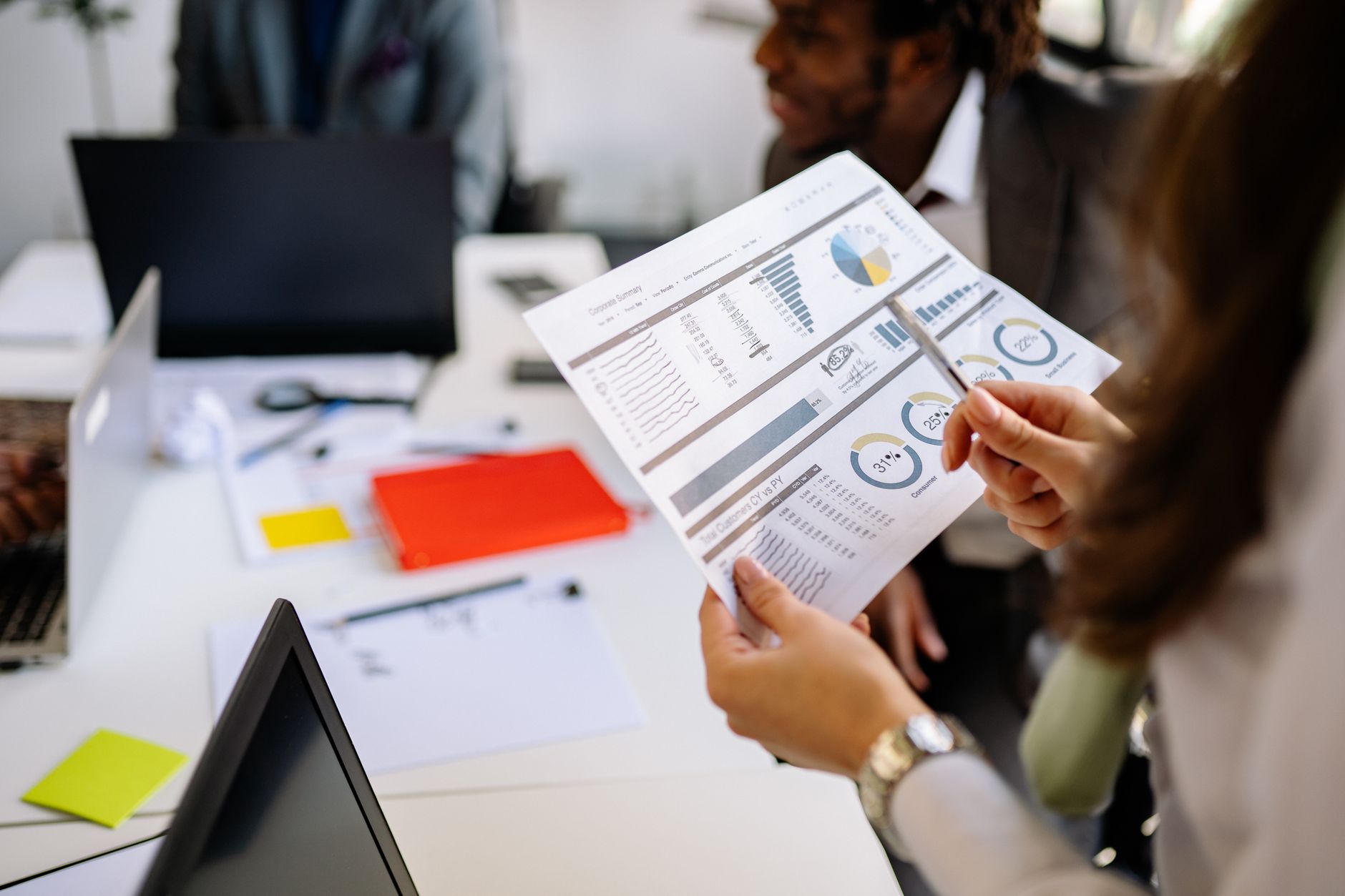 Une personne tient une feuille de graphiques et un stylo à une table de bureau avec ordinateurs portables, classeur rouge et collègue souriant en arrière-plan.