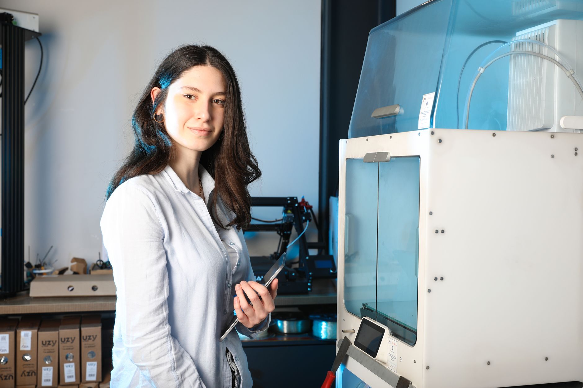 A woman in a light shirt holds a tablet in a workshop, standing next to a large enclosed 3D printer.
